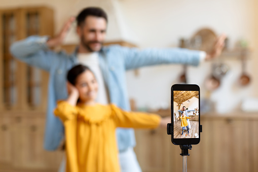 Social Media Fun. Cheerful Middle Eastern Father And Daughter Influencers Shooting Dance Video On Cellphone At Home, Creating Content for Family Online Vlog. Selective Focus On Smartphone Screen