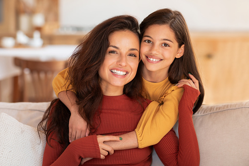 Mother's Love. Portrait Of Happy Young Arab Woman And Her Preteen Daughter Hugging Posing On Couch At Home, Smiling To Camera. Loving Mom Bonding With Kid Girl Embracing Indoors