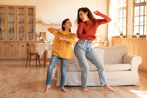 Positive Arab Young Mom And Preadolescent Daughter Dancing Together Standing In Modern Home Interior, Haivng Fun On Weekend. Kid And Mom Fooling During Domestic Party For Two, Full Length