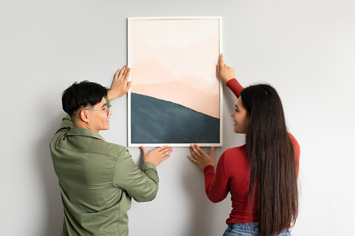 Interior Decoration. Japanese Young Husband And Wife Hanging Picture In Frame On Wall At Home, Back View. Married Asian Couple Decorating Their Living Room With Poster Together