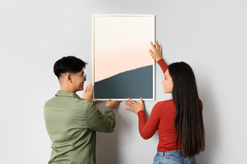 Chinese Young Spouses Decorating Their Living Room Wall With Modern Art, Hanging Poster In Frame Together Standing Indoors. Home Interior And Domestic Decoration Leisure Concept
