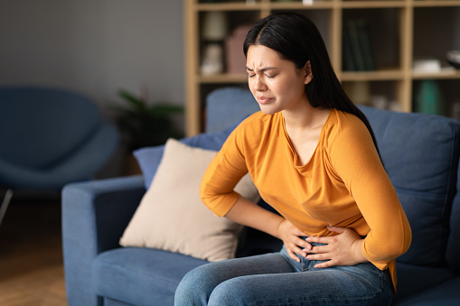 Painful Periods. Unhappy Asian Lady Struggling With Menstrual Cramps And Stomachache Pain, Touching Aching Belly Sitting On Sofa At Home. Female Health Problem, Stomach Diseases Concept