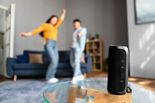 Wireless Smart Speaker Standing Playing Song For Unrecognizable Asian Couple While They Dancing And Having Fun, Listening To Favorite Music At Home. Shallow Depth, Focus On Musical Column Device
