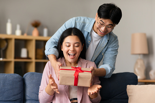Loving Korean Boyfriend Giving Gift Box To Girlfriend, Surprising Her On Birthday Holiday Sitting On Couch At Home. Family Celebrating Special Date, Anniversary. Presents And Celebration Concept