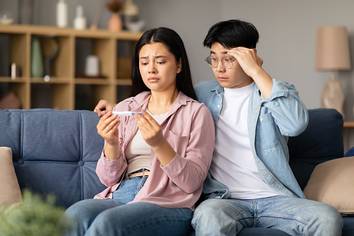 Unhappy Japanese couple holding a negative pregnancy test result, sitting on sofa, hugging and providing support amid this difficult moment at home. Reproductive and childbirth healthcare struggle