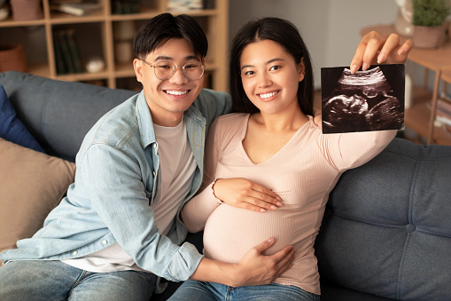 Joyful japanese couple showing sonogram photo of their baby embryo sitting on sofa indoor. Happy husband touching pregnant wife's belly while she holds ultrasound image announcing pregnancy