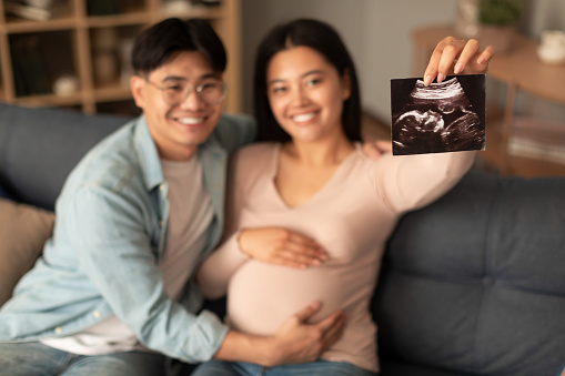 Cheerful pregnant korean young spouses showing ultrasound embryo photo, awaiting baby and announcing pregnancy sitting on sofa at home, smiling to camera. Selective focus on sonogram image