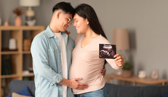 Awaiting Family. Loving japanese couple embracing, pregnant woman showing ultrasound photo of their baby embryo, standing at home interior. Selective focus on sonogram image