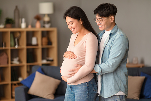 Pregnancy. Happy married japanese couple awaiting baby, standing together embracing in modern living room indoor, asian husband touching wife's belly. Family, joy of parenthood concept