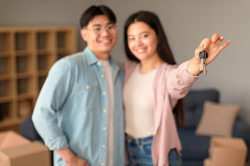 Real Estate Offer. Smiling Asian Spouses Stretching Their New House Keys To Camera At Home, Standing And Embracing Among Carton Moving Boxes, Ready For Move To A New Rental House. Shallow Depth