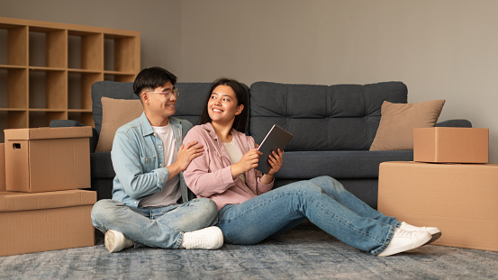 Young Japanese Couple Using Digital Tablet Sitting Among Moving Boxes On Floor In Modern Living Room, Searching Apartment For Rent Online At Home. Real Estate Website Advertisement. Panorama