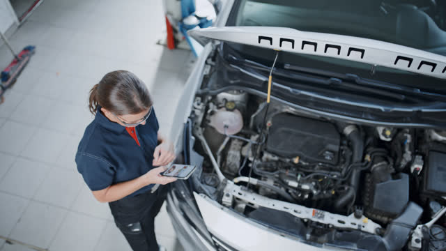 Female mechanic using smart phone at car engine in repair shop
