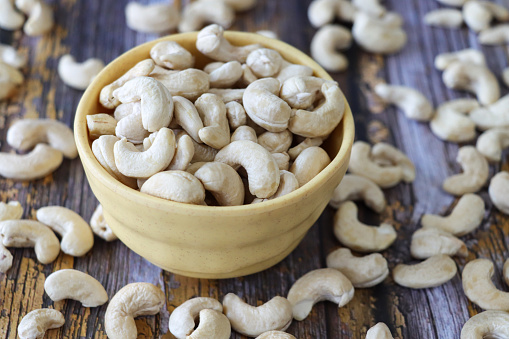 Stock photo showing close-up, elevated view of some cashews piled high in a yellow dish, against a woodgrain background. Raw cashews are actually unsafe to eat due to containing urushiol which is toxic and can cause skin irritation. Therefore, 'raw' cashews sold in stores have actually been cooked to remove the urushiol and once this is done cashews are considered to be a very healthy snack food being low in sugar but high in fibre as well as a good source of copper, magnesium, and manganese.