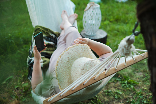 Weekend Vibes: A Hat-Clad Lady Makes the Most of a Sunny Afternoon, Engrossed in Her Phone While Swinging in Hammock