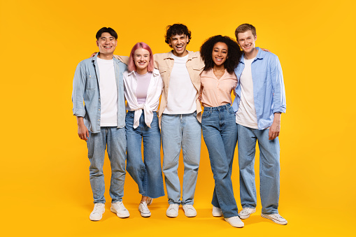 Group portrait of happy diverse friends hugging each other and smiling at camera, posing over yellow studio background, full length. Youth and friendship concept
