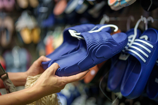 In the close-up shot, a woman is seen selecting sandals at an Asian street market stall. She appears to be carefully examining the options available to her, suggesting that she is looking for the perfect pair.