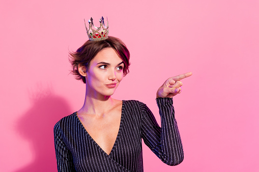 Photo of sweet unsure girl dressed glamour clothes tiara pointing empty space isolated pink color background.