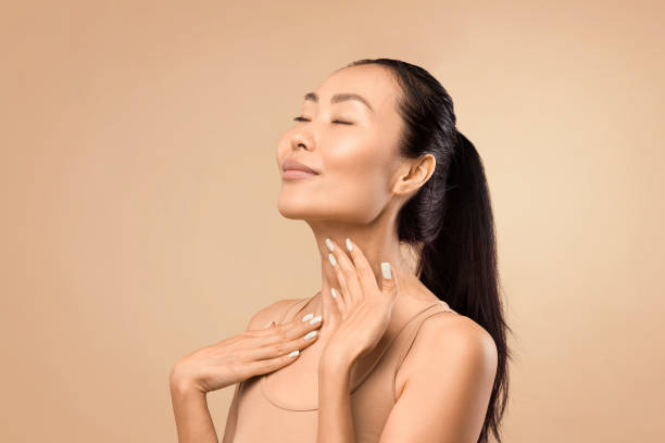 Middle-aged Asian woman touching smooth skin on neck, beige studio backdrop