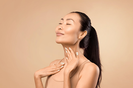 Middle-aged Asian woman with closed eyes gently touching her neck, expressing satisfaction with her skin's condition, studio shot with a beige background