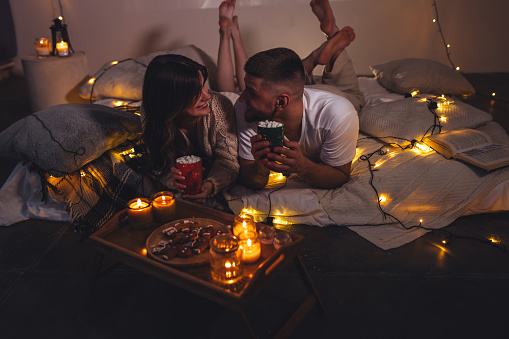 Cozy romantic evening, home weekend In bed for a loving couple. Candles, movies, hot chocolate with marshmallow, popcorn. Beautiful young man and woman enjoying relationship, having fun, laughing
