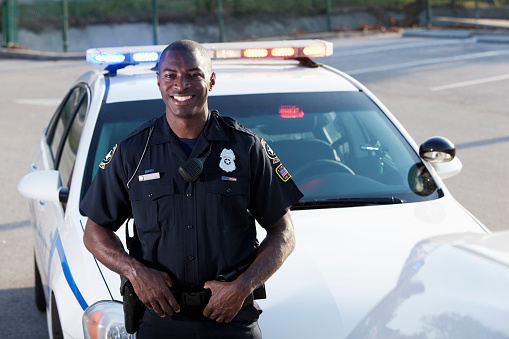 African American police officer (20s) standing in front of cruiser.