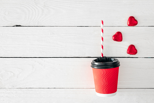 Red paper cup and heart-shaped candies on a white wooden background. View from above. Space for text. Valentine's Day concept.