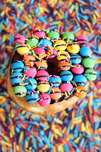 Stock photo showing close-up, elevated view of a glazed doughnut with a hole, decorated with chocolate drops covered with a hard candy coating, on a bed of rainbow coloured sugar strand sprinkles used for the decoration of cakes.