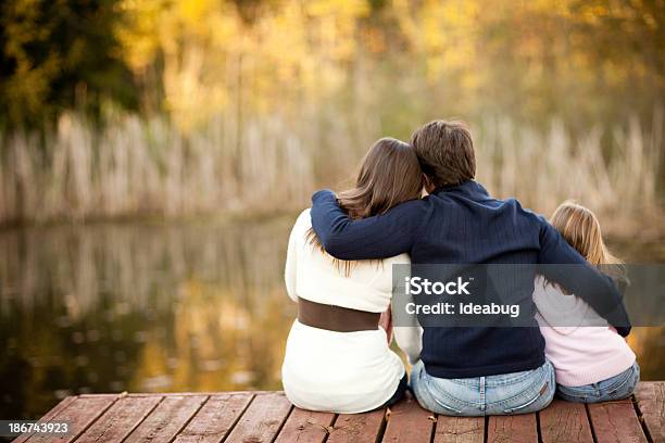 Family Of Three Sitting On Deck Overlooking Pond In Autumn Stock Photo - Download Image Now