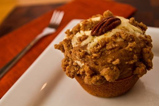 Delicious homemade cheescake muffin with frsoting under apple crumble and pecan on top resting on a white plate, evoking comfort and indulgence. Perfect for bakeries and recipe websites.