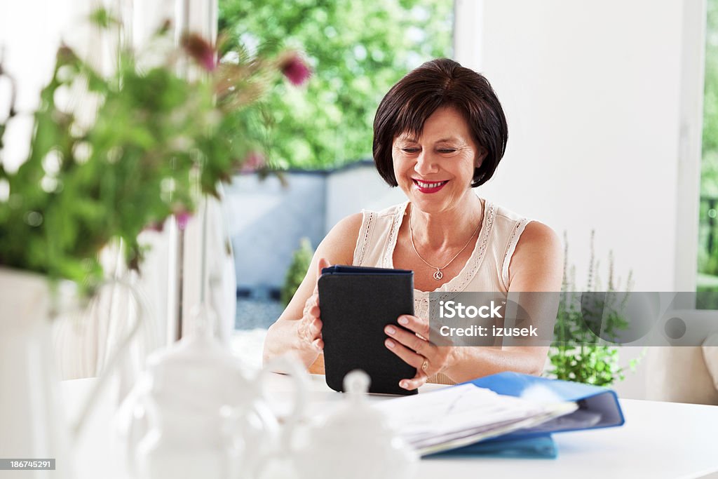 Mature woman with e-reader Cheerful senior woman sitting at the table at home and using an e-reader. 55-59 Years Stock Photo Mature woman with e-reader Cheerful senior woman sitting at the table at home and using an e-reader. 55-59 Years Stock Photo