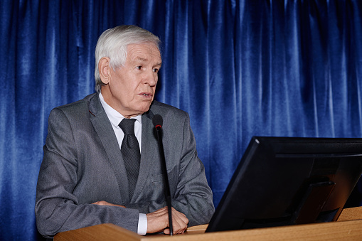 Mature businessman speaking in microphone and presenting his report at conference