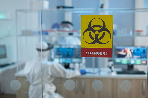 Selective focus on danger sign in microbiological hospital laboratory during chemical experiment. Teamwork of scientist with coronavirus protective medical equipment working at medicine vaccine