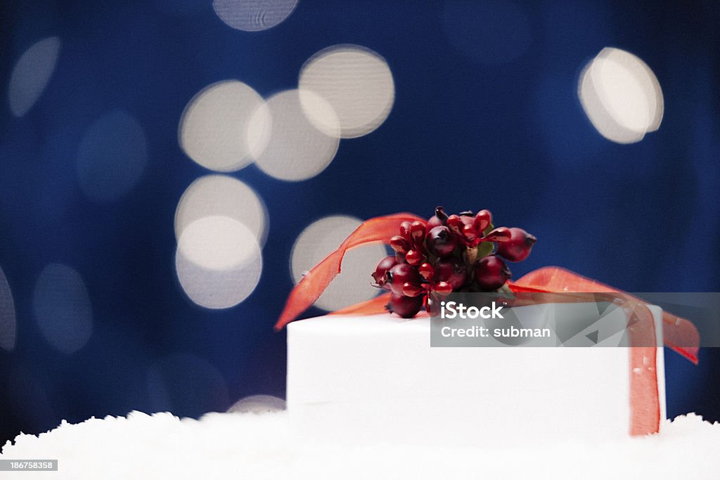 Christmas Git In The Snow A wrapped Xmas gift with red ribbon & a sprig of holly lies in the snow, twinkly lights on a blue background Blue Stock Photo