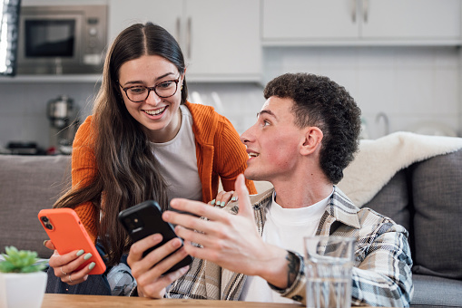 A medium front view of two young students sitting in the common room they are smiling and comparing their lectures on their mobile phone. They are enjoying a break between lectures and lounging together in the shared social space of their university.