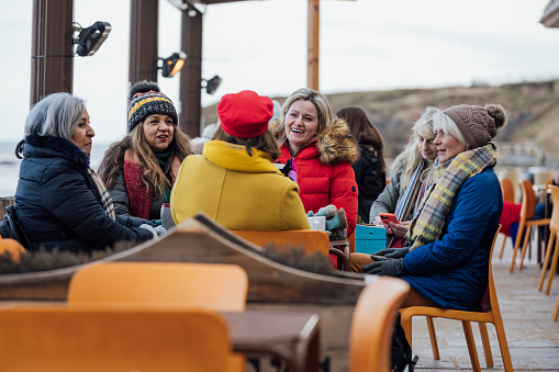 A group of mature female friends sitting outside at a bar/coffee shop in Tynemouth, North East England. They are wearing warm clothing to keep warm in January while they sit and chat with each other during a catch-up.