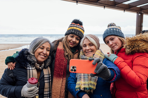 A group of mature female friends wearing warm clothing, standing outdoors and spending the day together at the beach in Tynemouth, North East England. They are taking a selfie together on a mobile phone.