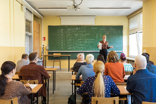 Back view of group of mature adult female and male students sitting in the classroom. Female teacher standing in front of blackboard.