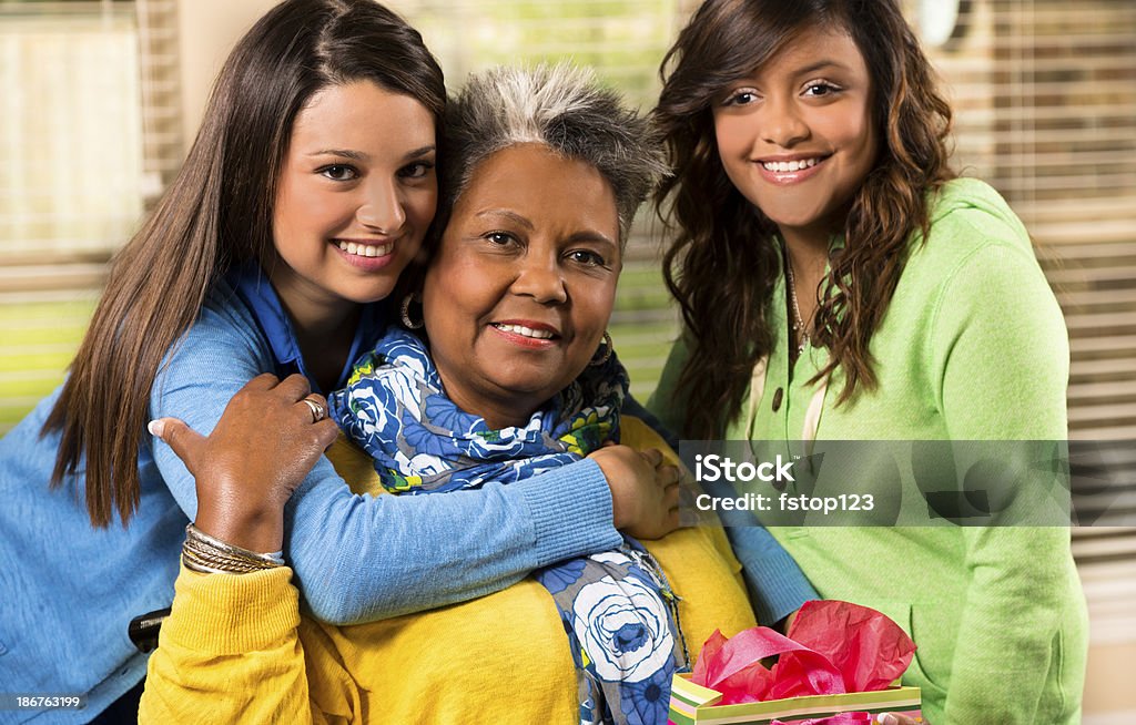 Young women smile and pose with senior woman Two young ladies visiting an older woman in her home.  The girl on the left hugs the senior while her friend watches.  The two ladies and the senior are smiling and facing the camera.  The girl on the left is wearing a blue sweater.  The senior has gray hair and is wearing a yellow jumper and a blue flower-patterned scarf.  She is also wearing silver bangles and holding a red flower.  The girl on the right is wearing a green jumper.  The wall, door and glass window appears in the background. Family Stock Photo