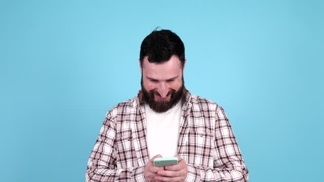 Young bearded man looking at phone and typing, standing isolated on blue background.