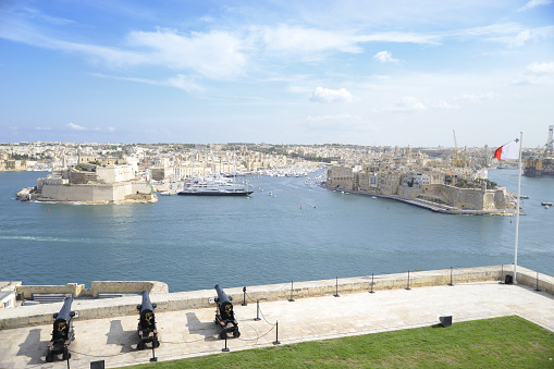 A scenic view of Valletta's historic fortified city and bustling harbor under a clear blue sky, with cannons in the foreground as a nod to its storied past