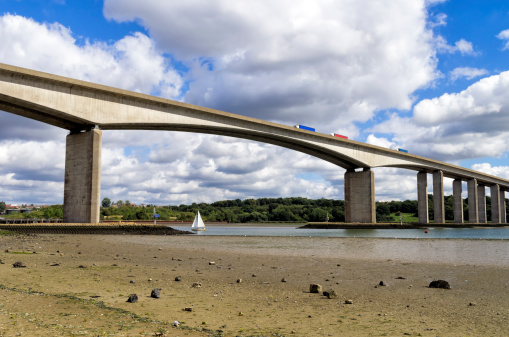 A small dinghy sailing upstream below the Orwell Bridge, which crosses the River Orwell at Wherstead, near Ipswich in Suffolk, England. Three lorries are on the bridge.