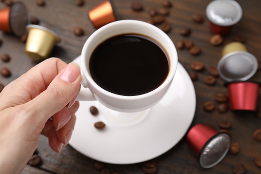 Woman holding cup of coffee at wooden table with capsules and beans, closeup