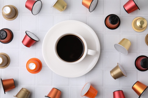 Cup of coffee and capsules on white tiled table, flat lay