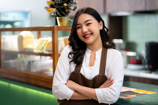 Smiling young asian waitress wearing an apron and smiling while standing in a restaurant