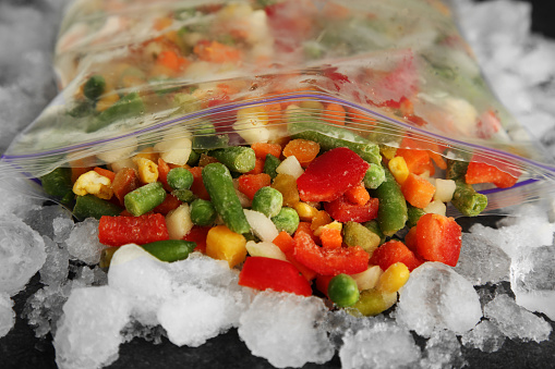 Zip bag with different frozen vegetables and ice on table, closeup