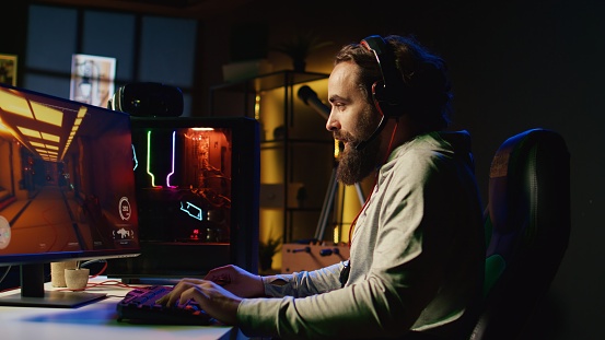 Happy man putting on headphones to communicate with teammates online from his dark living room, playing engaging video games on gaming PC at computer desk, chilling after work