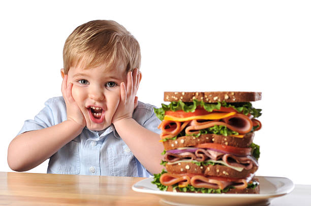 Young Boy with Triple Decker Dagwood Sandwich cute image of a little boy with a very large sandwich. It is layered with deli meats, cheeses, purple onion, tomato and lettuce on wheat bread. The boy has a very surprised look on his face. How will he be able to eat all of it? dagwood stock pictures, royalty-free photos & images