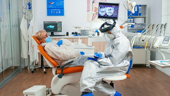 Dentist assistant in coverall talking with senior woman during global coronavirus pandemic writing on clipboard. Nurse and orthodontic doctor wearing face shield, protection suit, mask and gloves.