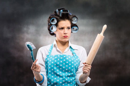 Portrait of frustrated housewife wearing blue apron and curlers, holding roller pin and a brush.