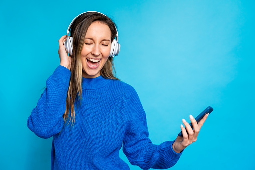 Happy young woman listening to music with headphones and smartphone on blue background